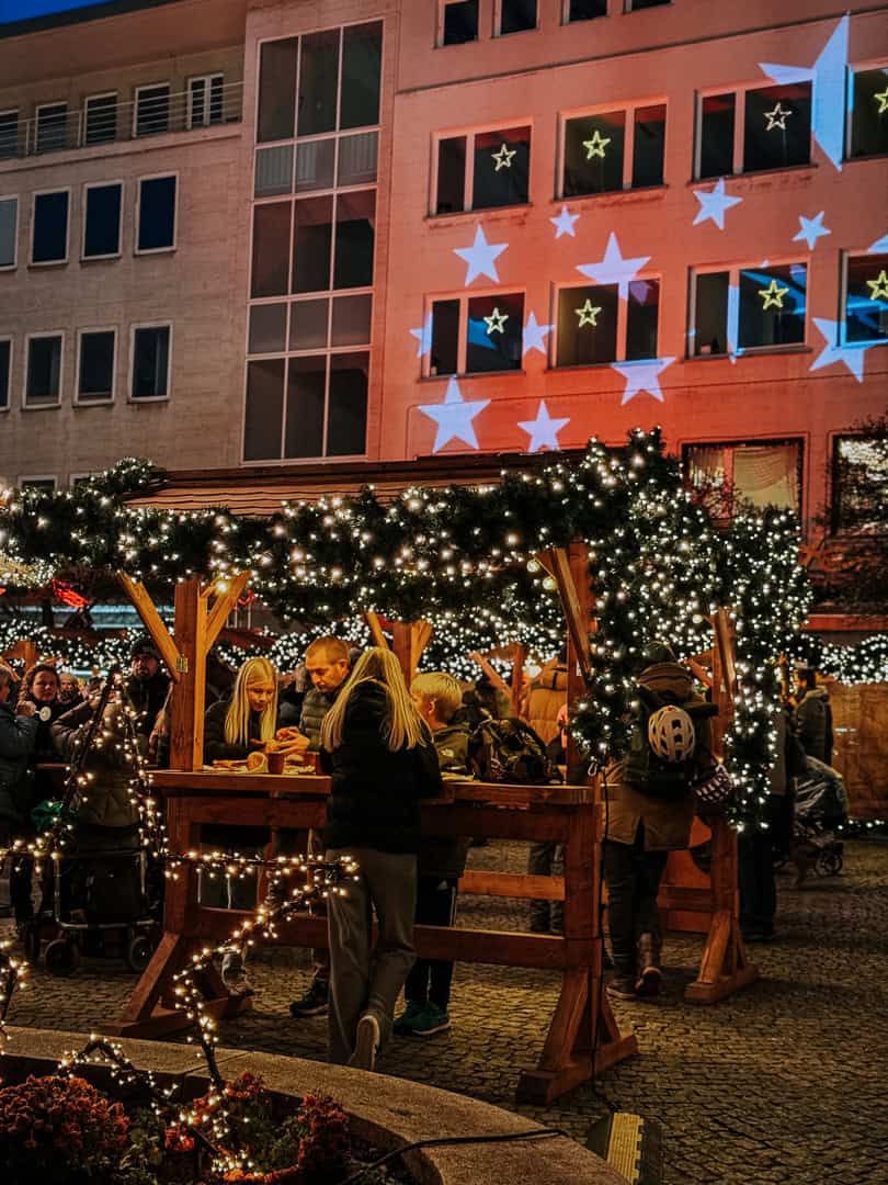 A wooden market stall illuminated with string lights serves as a gathering spot for families enjoying festive treats under a sky filled with projected blue stars.