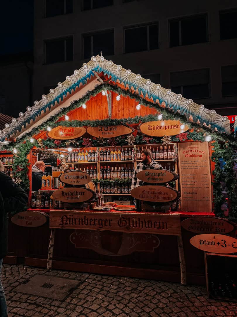 A charming Christmas market stall adorned with festive decorations offers a selection of mulled wine, as indicated by the 'Nürnberger Glühwein' sign, inviting passersby to enjoy the holiday spirits.