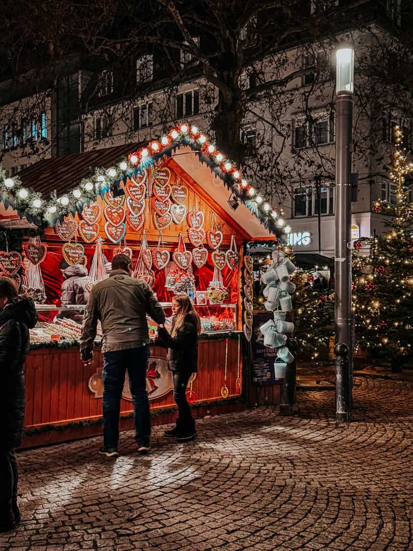 Customers browse an array of classic gingerbread hearts at a warmly lit Christmas market stall, adding to the festive spirit with its traditional sweets and treats.