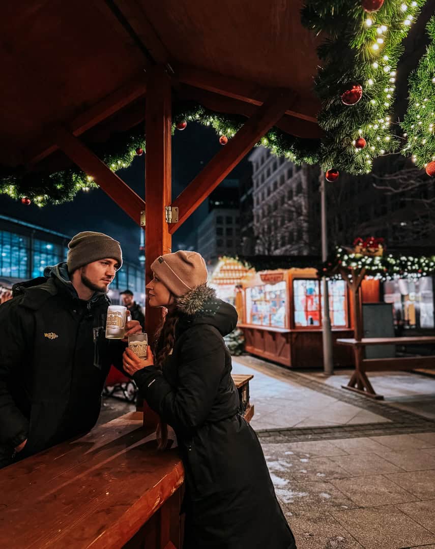 A couple stands close, sharing a moment and a mug of glühwein under a gazebo adorned with Christmas lights, with the bustling market and its visitors in the background.