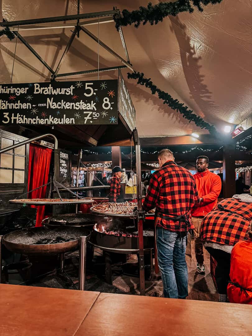 A bustling Christmas market booth in Berlin offering traditional grilled sausages and meats, with a visible price list and festive decor.