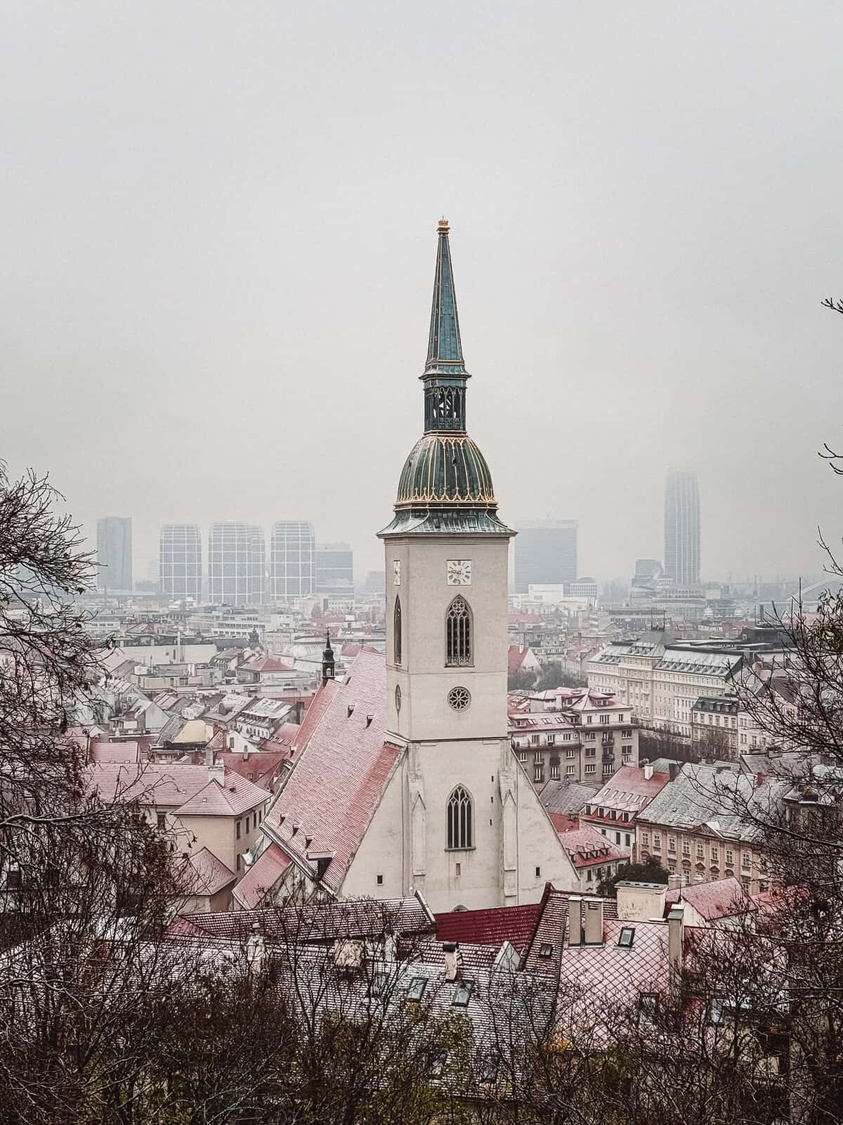A view of St. Martin's Cathedral spire rising above the surrounding historical architecture in Bratislava, contrasting modern buildings in the hazy background, showcasing the city's blend of old and new.