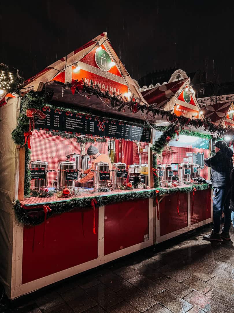 Another angle of a festive outdoor market stall adorned with Christmas decorations selling various hot drinks, with prices listed in euros, as customers gather around.