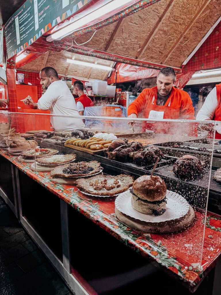 A lively food stall at a Christmas market, where vendors in red aprons grill meats and serve burgers and pastries to eager customers, with the stall front covered by a protective clear screen.