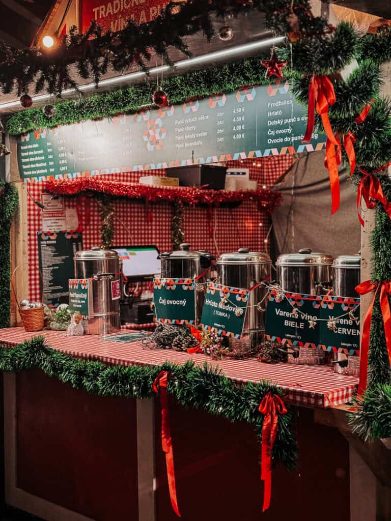 : A cozy market booth selling hot drinks for chilly evenings, decorated with red bows and garlands, featuring a festive banner with drink options and prices in euros.