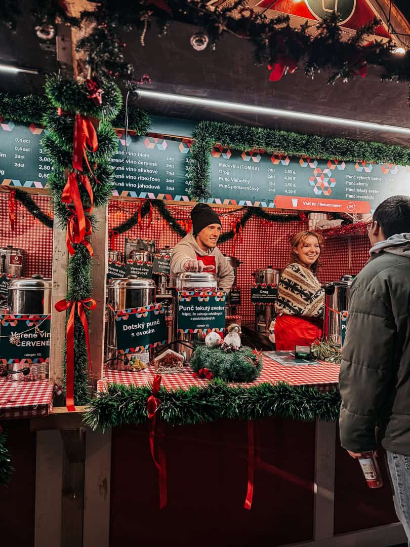 A Christmas market booth with a vendor in a beanie serving drinks and interacting with customers; the stall is adorned with greenery and festive decorations, listing drink names and prices.