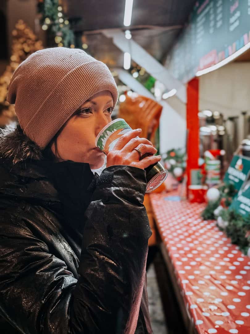 A close-up of a woman in a beige beanie sipping a hot drink from a festive cup, with the Christmas market lights softly blurred in the background.