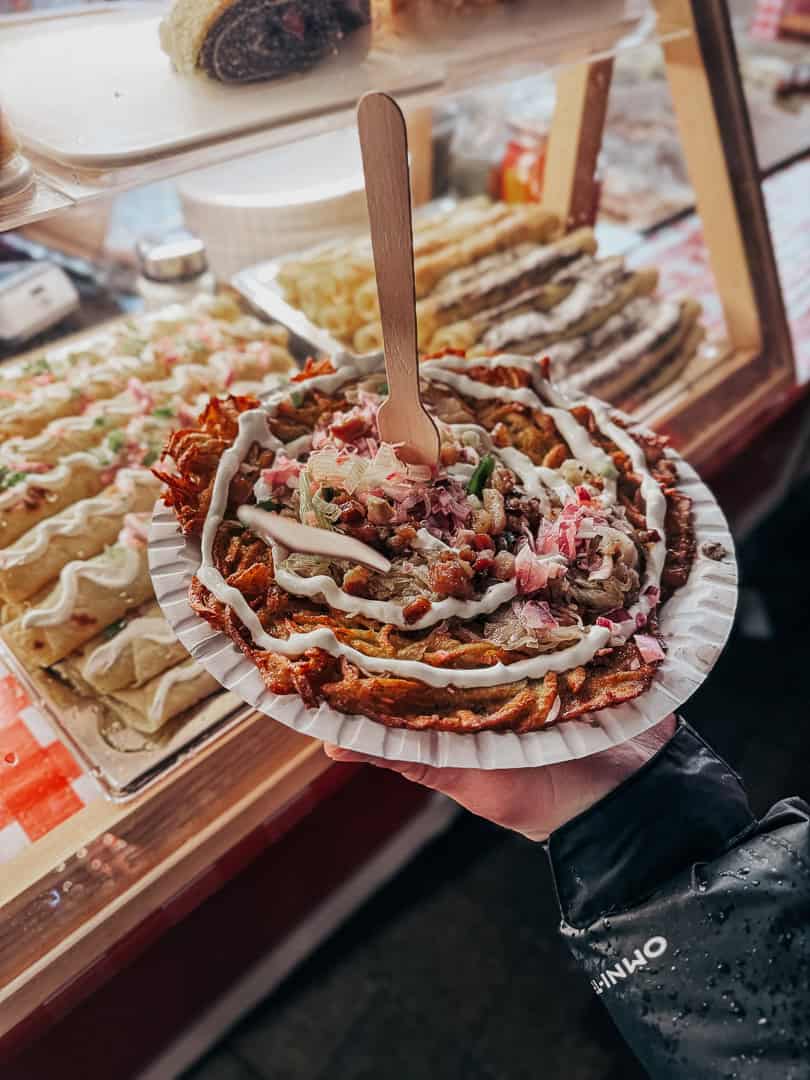 A hand holding a paper plate with a freshly made street food item, topped with a variety of condiments and a wooden fork stuck in the middle, with more food items visible in the background on a street food stall