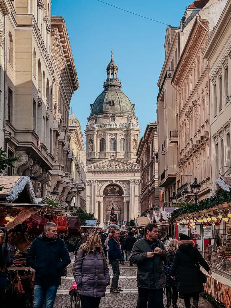 A bustling Christmas market lane leading towards a magnificent, historic domed building, with shoppers browsing the festive stalls under a clear blue sky.