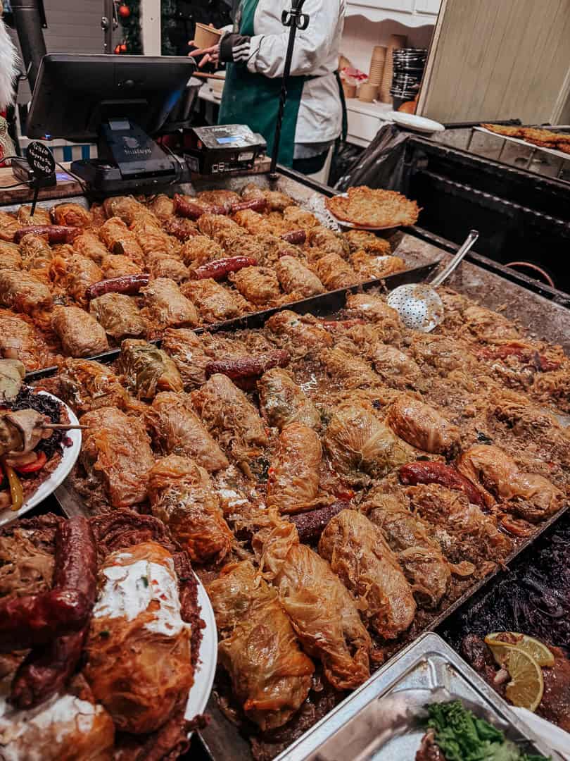 A bustling street food stall with a variety of stuffed cabbage rolls and sausages served on a large grill, surrounded by eager customers waiting to be served.