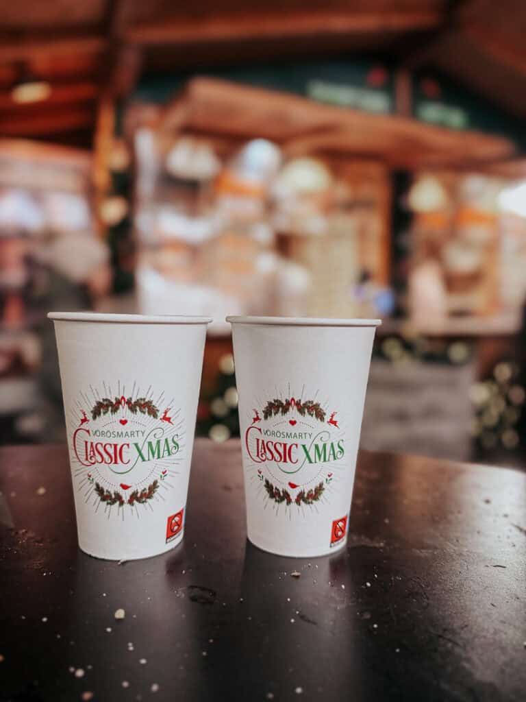 Two cups of 'Vörösmarty Classic Xmas' mulled wine sit on a table with a festive blurry background, highlighting the holiday market's offerings.
