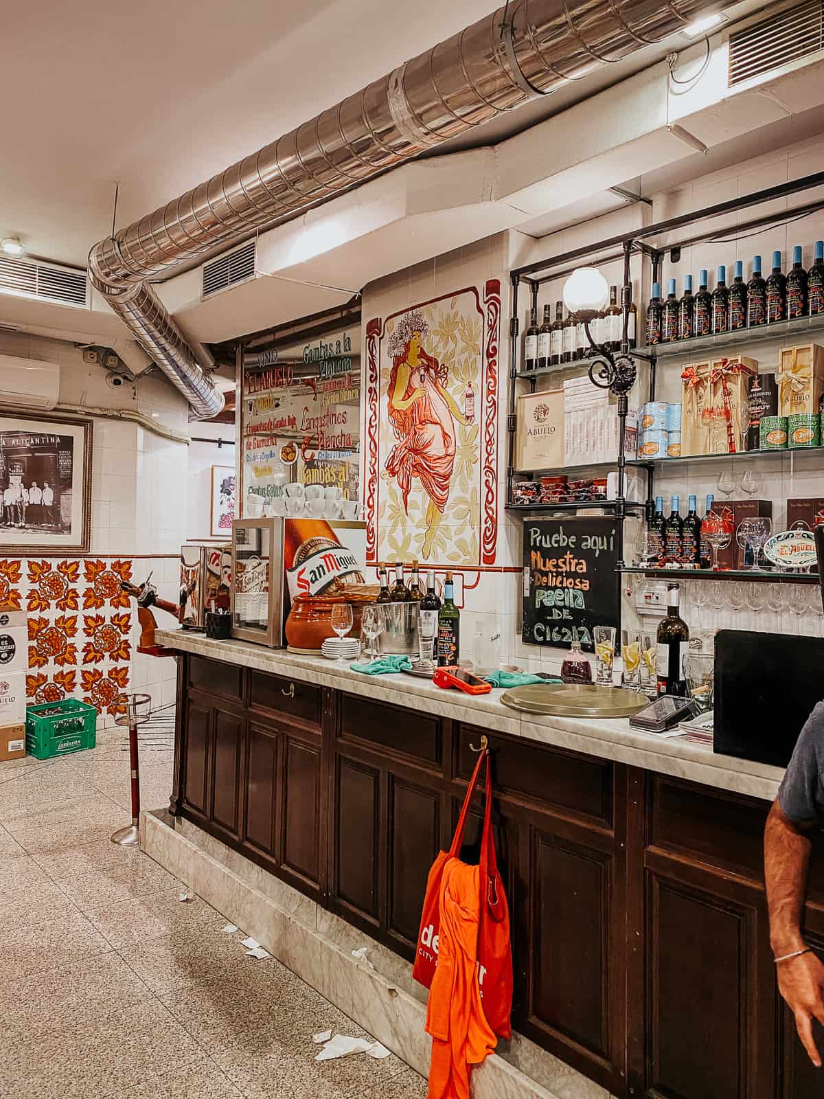 A vibrant Spanish tapas bar with traditional tiles and a large illustrated poster, showcasing a variety of local spirits and decorative red accents.