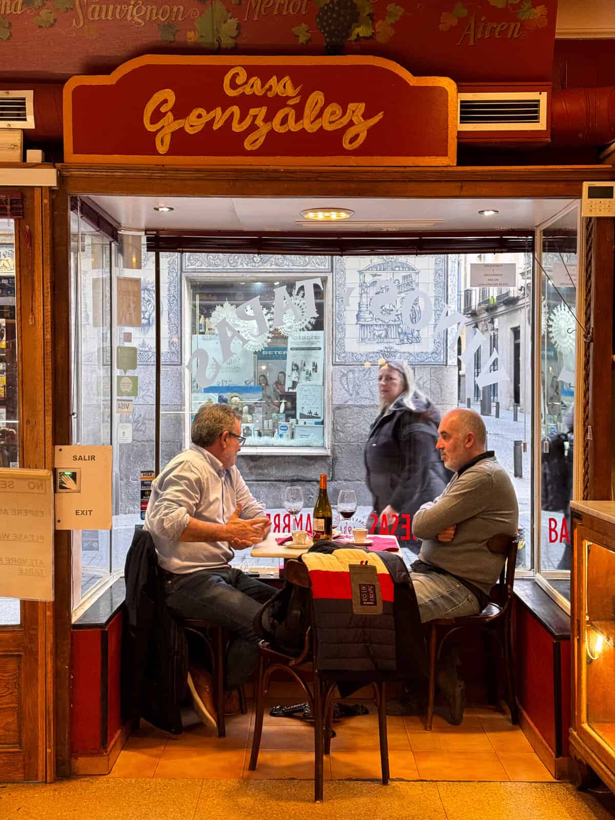 Two men engaged in conversation at a table inside Casa Gonzales, with wine and tapas between them, as a passerby blurs in motion outside the quaint window-front.