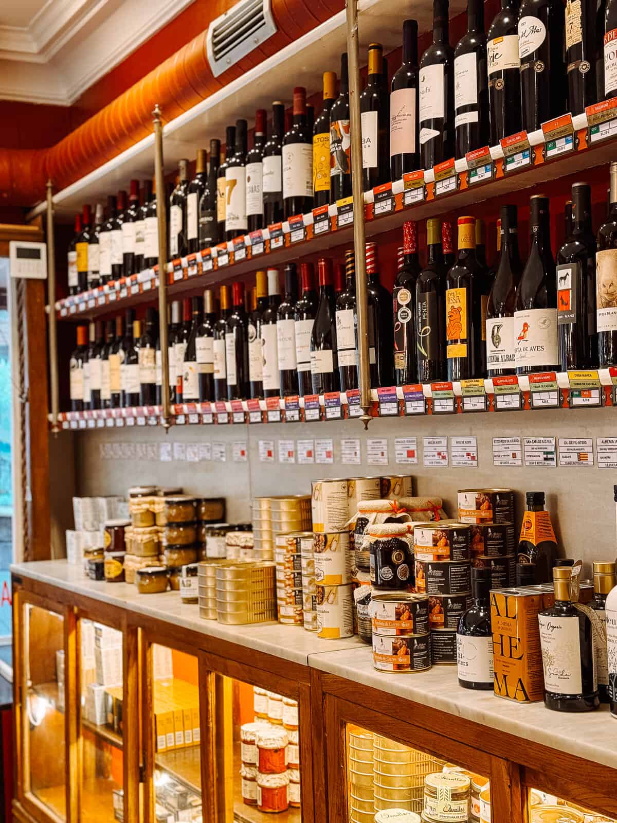 A well-stocked shelf in a gourmet shop featuring an assortment of red wines above and various tins of cookies and boxes of cheese below