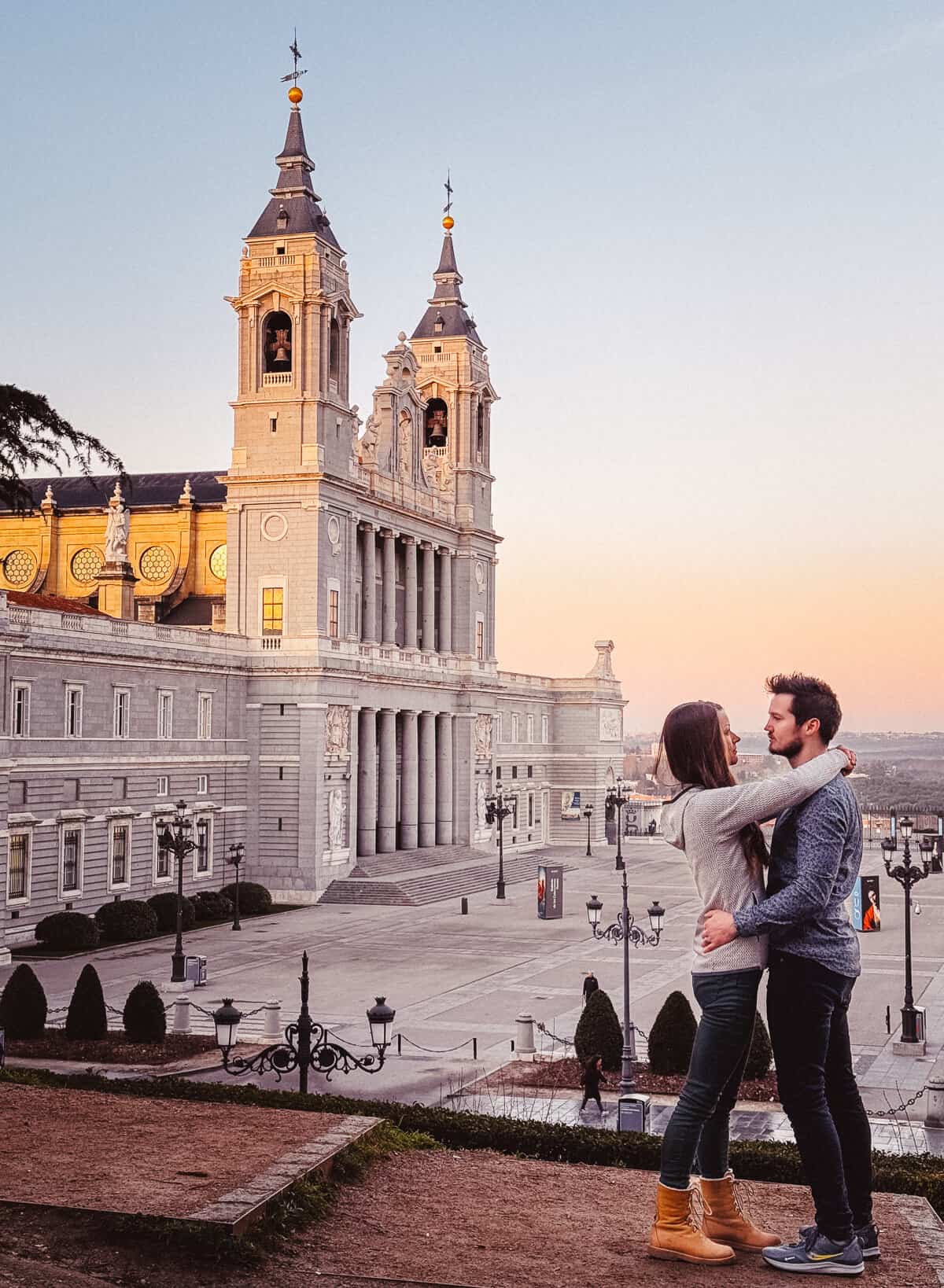 An intimate moment between a couple in front of the Almudena Cathedral in Madrid at dusk, with the historic structure illuminated, providing a majestic setting for a romantic scene.