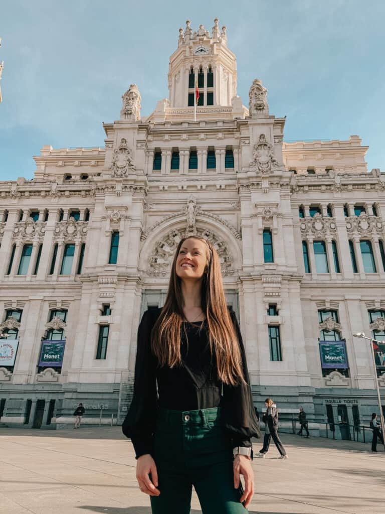 A smiling woman standing in front of the grand Cibeles Palace in Madrid, her cheerful presence complementing the elegance of the historic building bathed in sunlight.