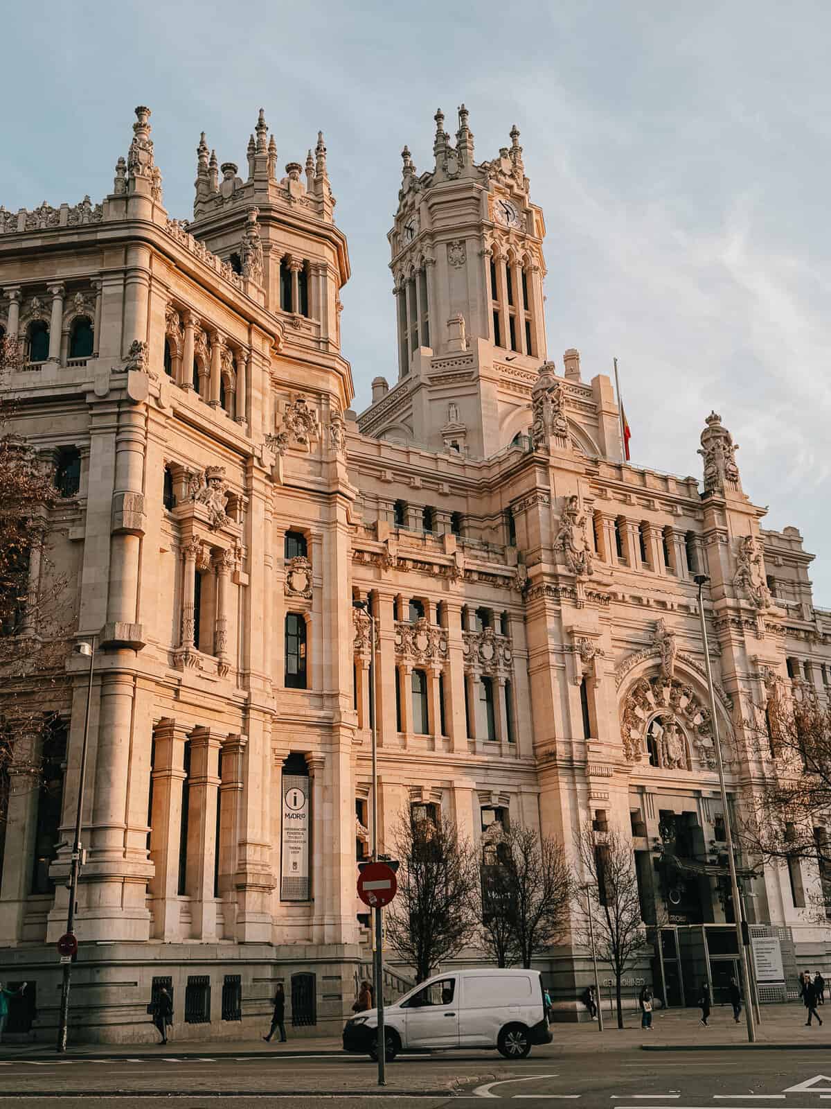 Cibeles Palace in Madrid, captured in soft evening light that casts a warm glow on the building's elaborate façade and detailed sculptures, with clear skies above and city life moving along its base.