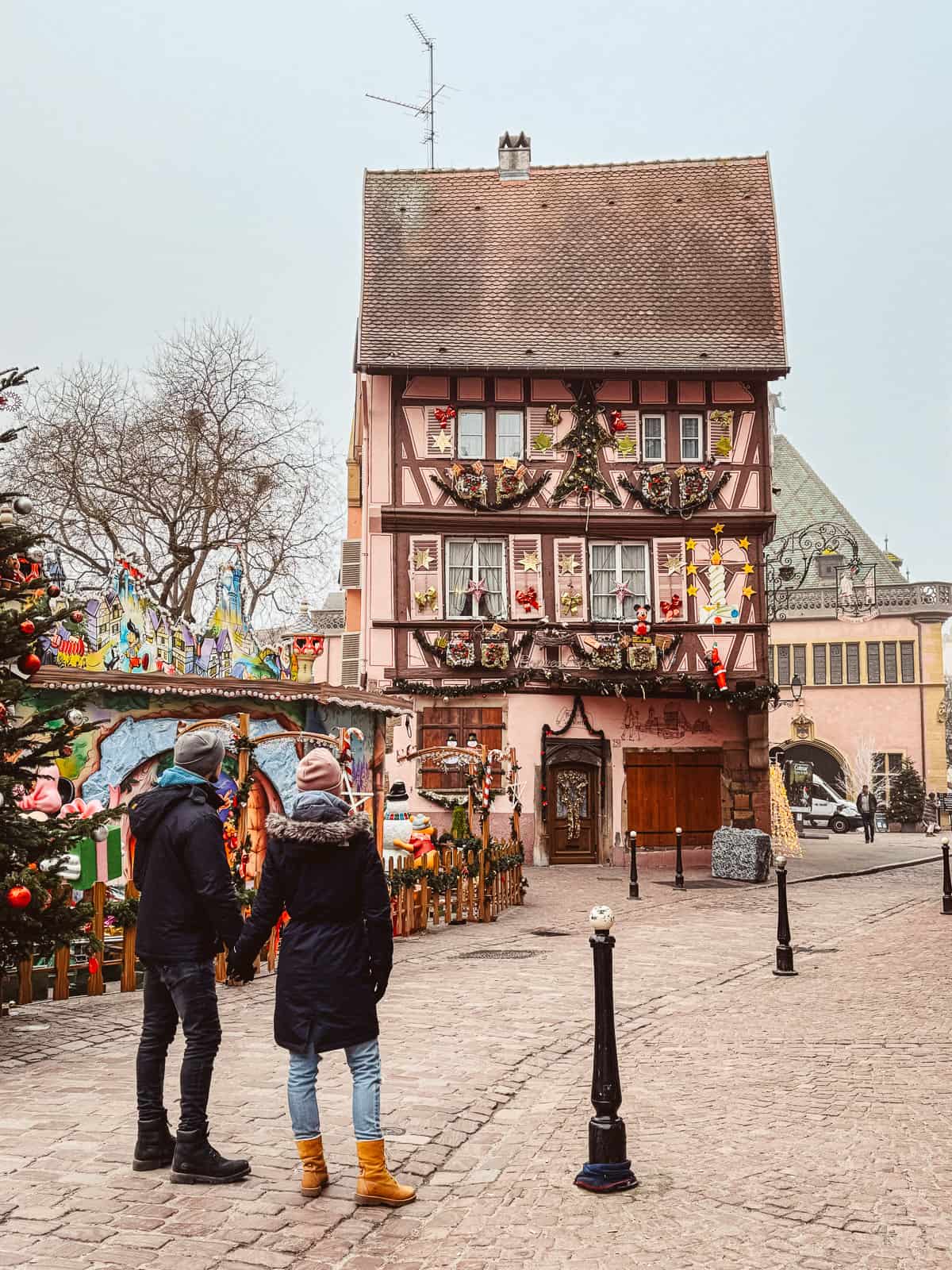 A couple stands admiring a whimsical pink half-timbered house adorned with Christmas decorations in Colmar, with a vibrant carousel and festive tree adding to the holiday spirit.