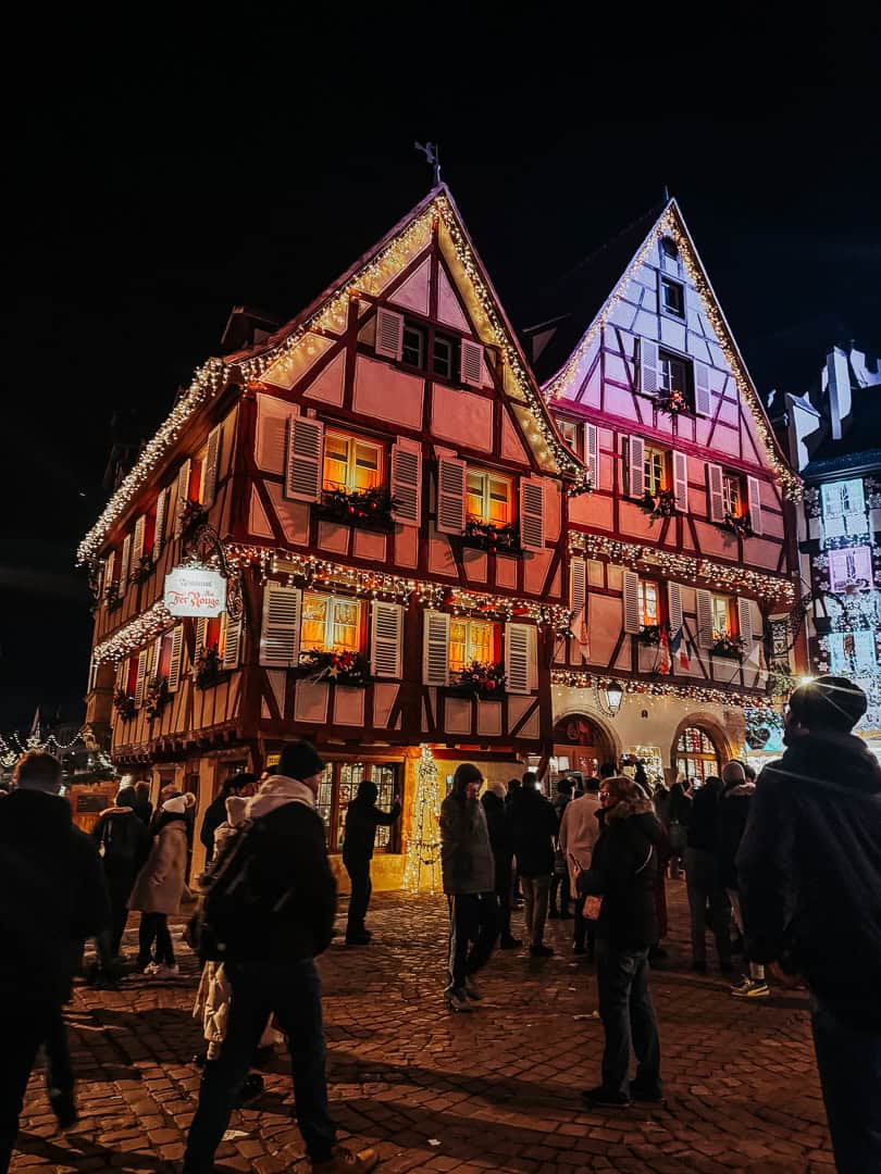 The warm, rich colors of historic half-timbered houses in Colmar are illuminated by festive lights at night, with a bustling Christmas market adding to the lively atmosphere.