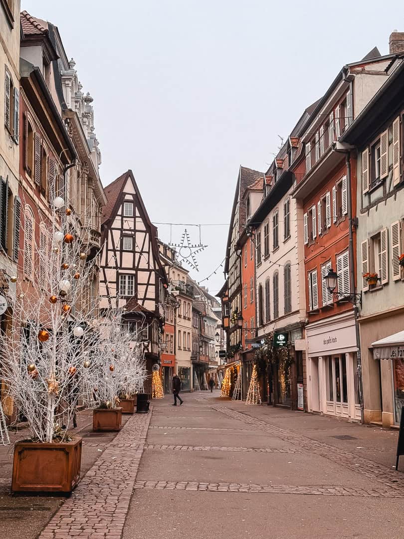 Deserted cobblestone street in Colmar decorated with Christmas lights and white tree ornaments, leading to an intricate wrought-iron street lamp and timbered houses.