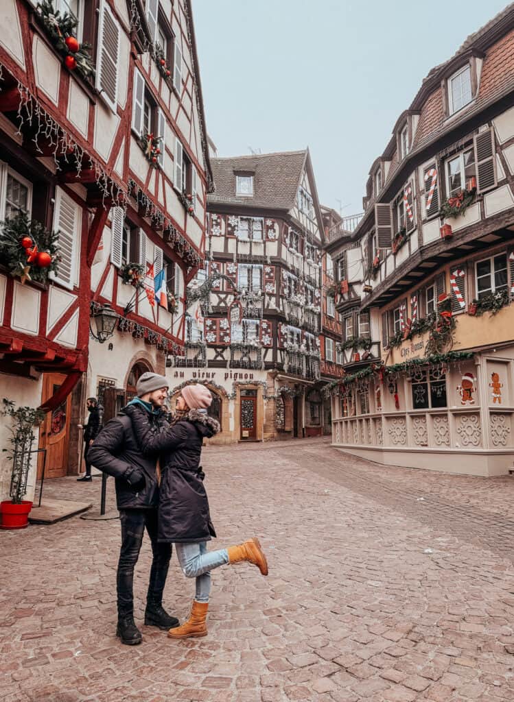 A couple embracing in a romantic moment on a cobbled street in Colmar, surrounded by charming half-timbered houses adorned with Christmas decorations.