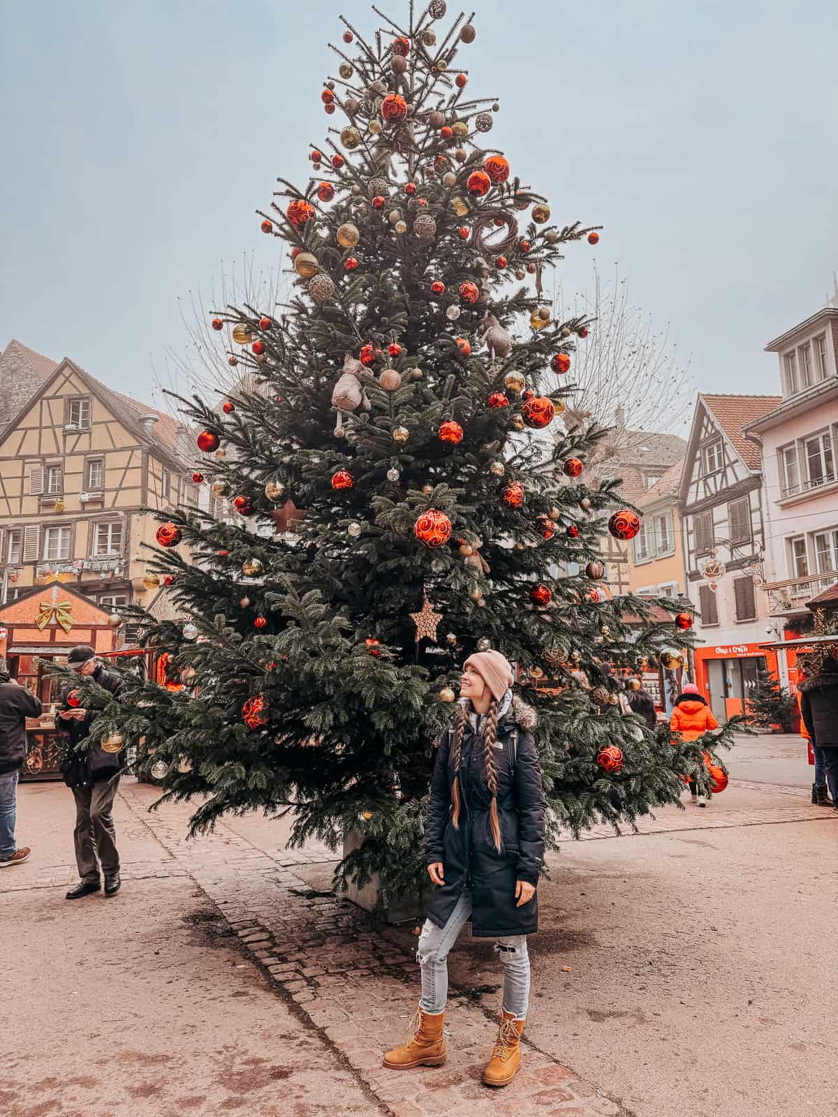 A person stands in awe before a towering Christmas tree beautifully decorated with red and gold ornaments in the festive streets of Colmar.