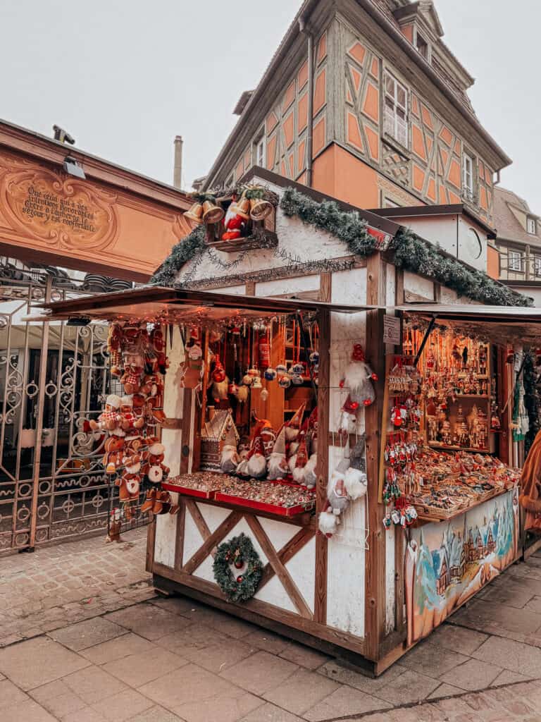 A quaint Christmas market stall in Colmar brimming with Santa figures, wreaths, and an array of red and white decorations, with half-timbered facades in the background.