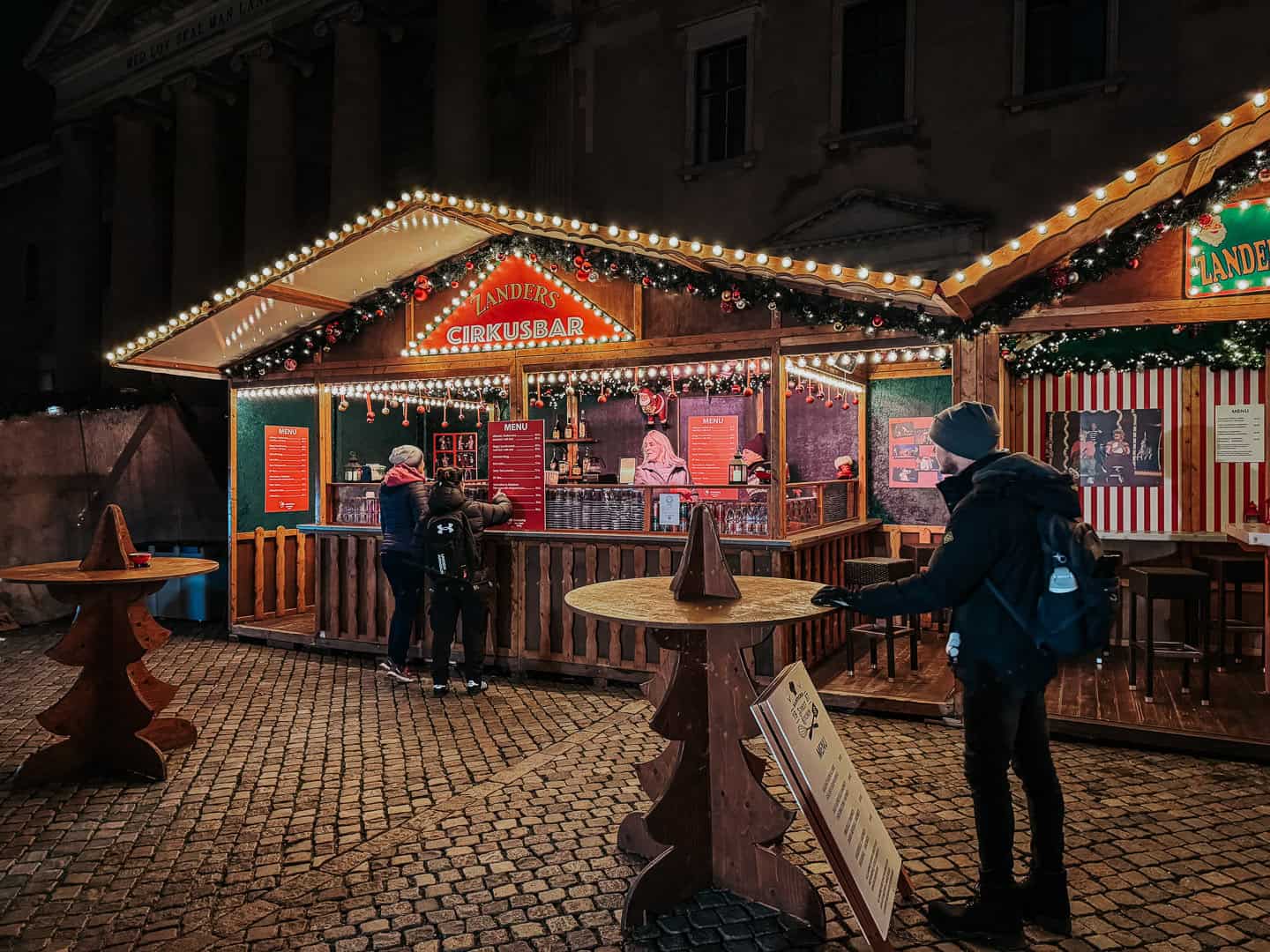 Nighttime at a Christmas market in Copenhagen with visitors gathered around Landers Cirkusbar, featuring a brightly lit menu and festive decorations
