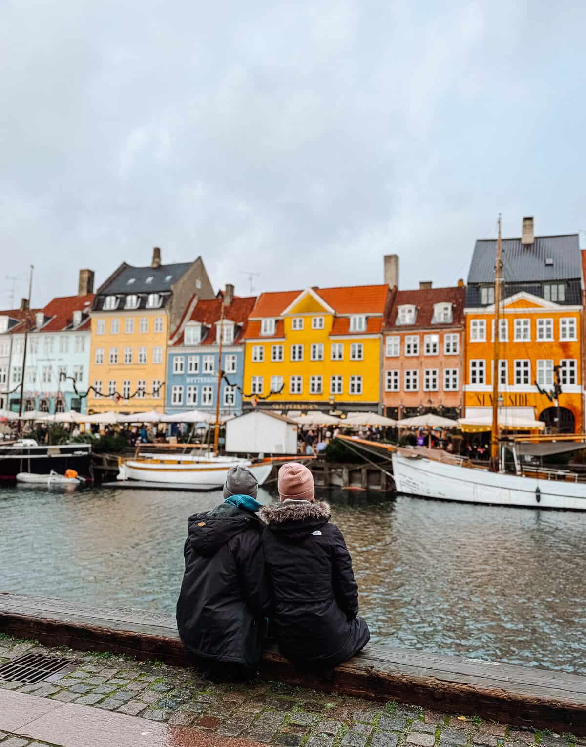 A couple sits by the canal in Copenhagen, facing the row of brightly colored buildings, enjoying the view despite the overcast sky.