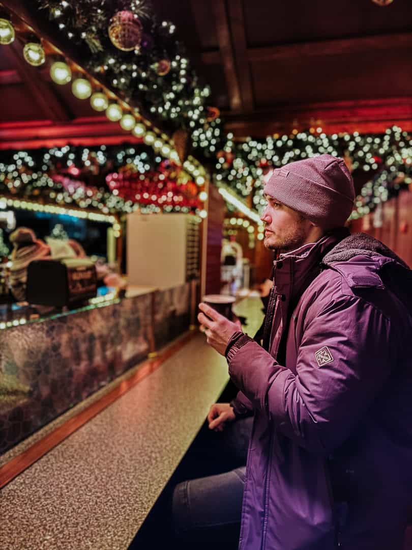A man in a purple jacket enjoying a hot beverage at a Christmas market, with twinkling lights and holiday decorations enhancing the festive mood.