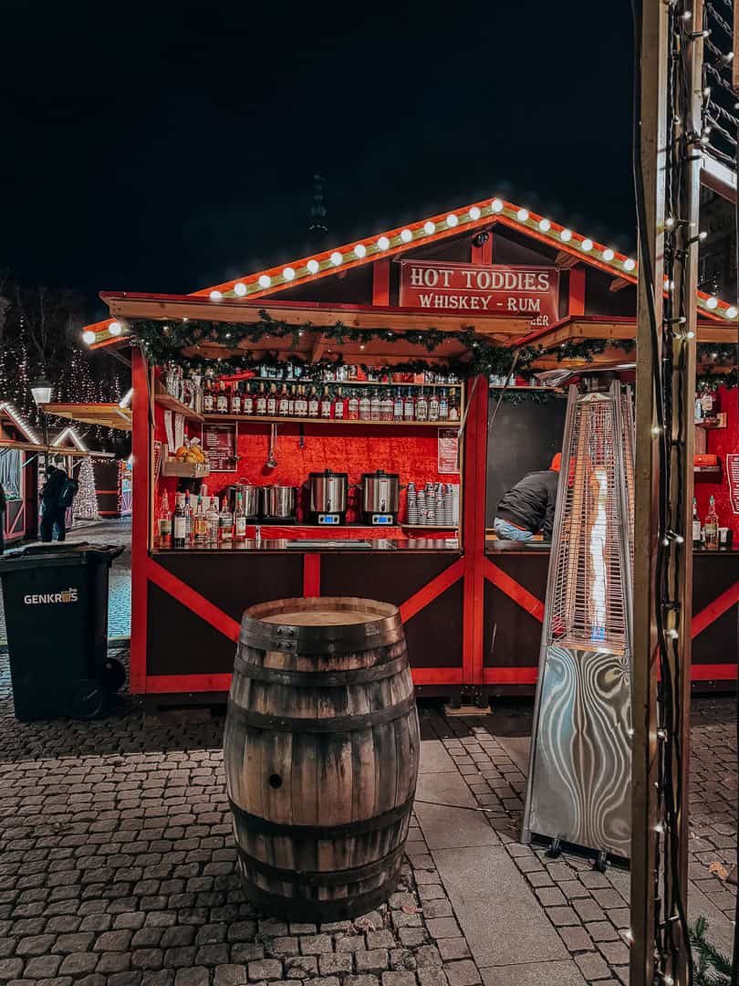 A Christmas market stall in Copenhagen illuminated at night, offering 'Hot Toddies Whiskey - Rum,' with a wooden barrel in the foreground set against the cobblestone street