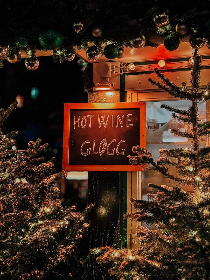 Close-up of a sign reading 'Hot Wine Gløgg' at a Copenhagen Christmas market, with a backdrop of festive greenery and shimmering baubles