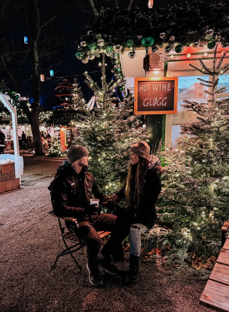 A couple enjoys a moment over hot drinks at a Copenhagen Christmas market, seated by a tree adorned with lights, giving a cozy and intimate atmosphere