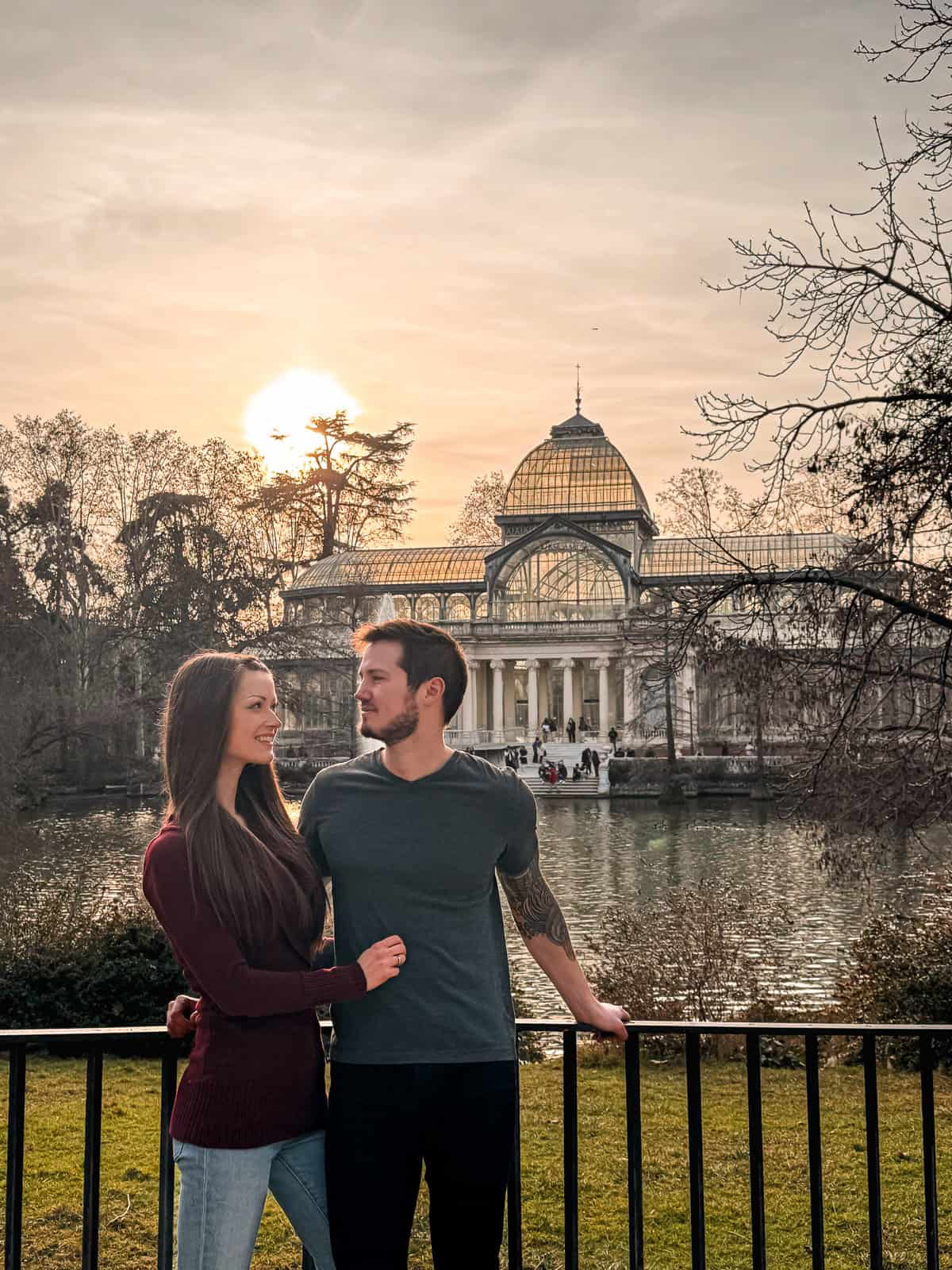 A couple stands in a loving embrace before the Crystal Palace in Madrid's Retiro Park, with the sun setting behind the glass structure, reflecting a golden hue over the peaceful scene.