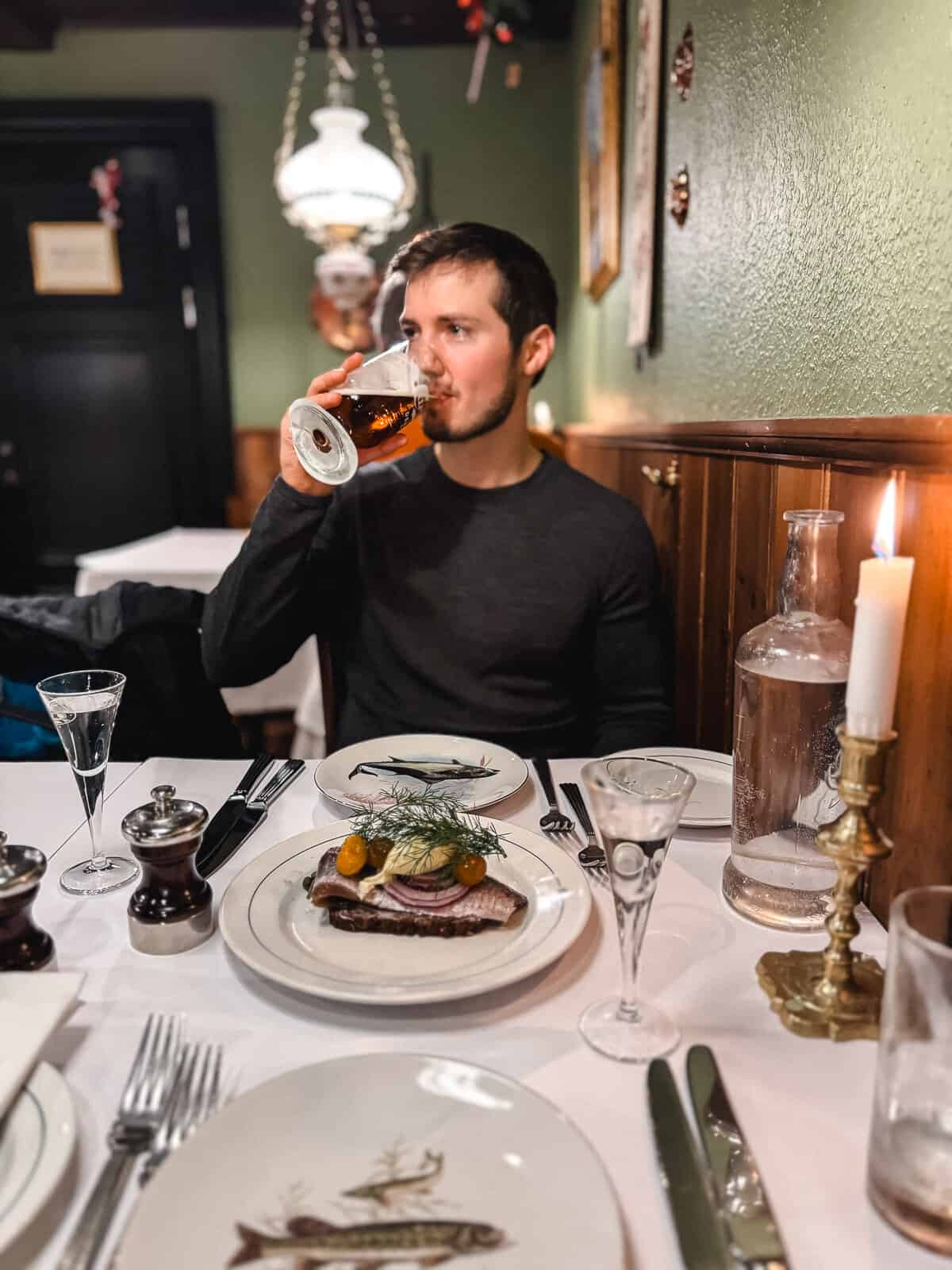 A man enjoys a beer in a cozy, traditional European restaurant, with a focus on a plate of delicately arranged herring, onions, and herbs in front of him, surrounded by elegant tableware and a romantic candlelit setting.