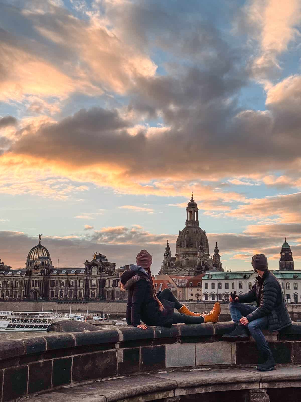 Two people sit on a wall overlooking the Dresden cityscape under a stunning sunset sky, with the iconic Frauenkirche and other baroque architecture in the background.
