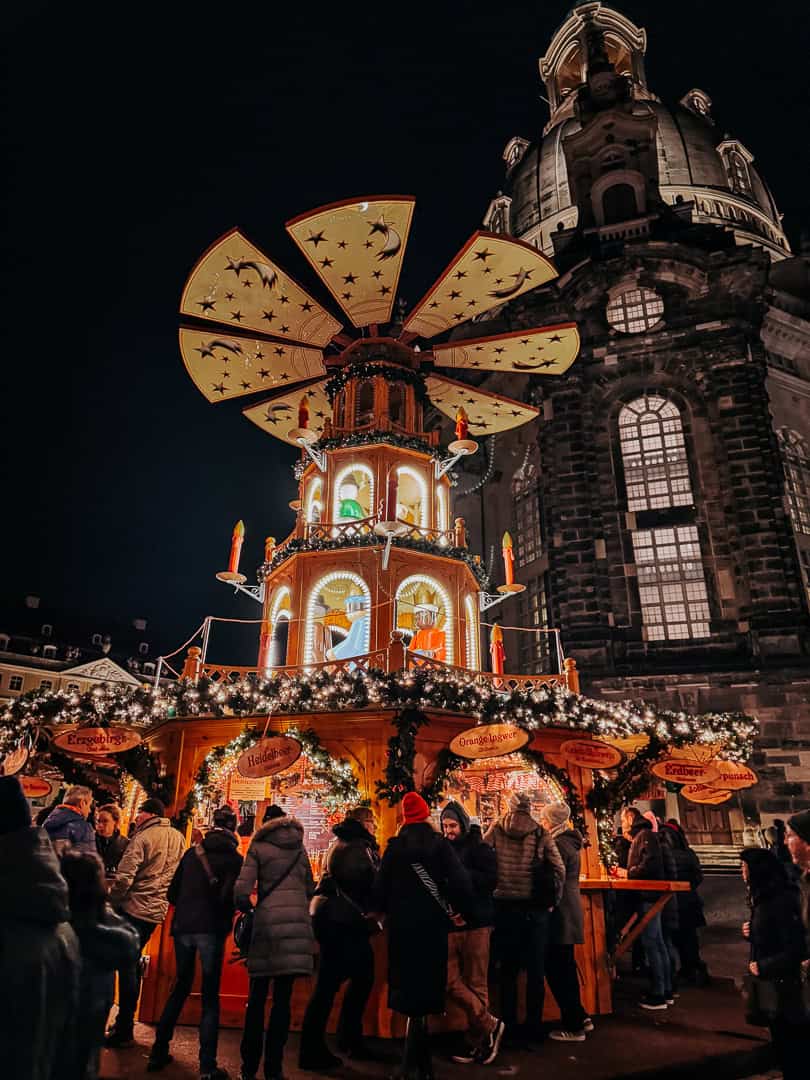 Visitors gathered around an elaborate Christmas pyramid decoration with lit candles, set against the historic backdrop of Dresden's architecture at night
