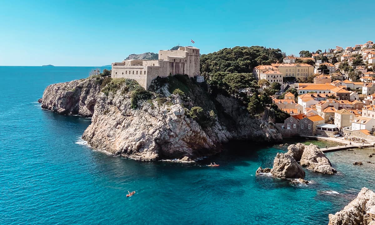 Aerial view of the coastal city of Dubrovnik, featuring the historic fortress perched on the edge of a cliff overlooking the Adriatic Sea, with the old town's orange-roofed buildings in the background and a few kayaks dotting the clear blue waters.