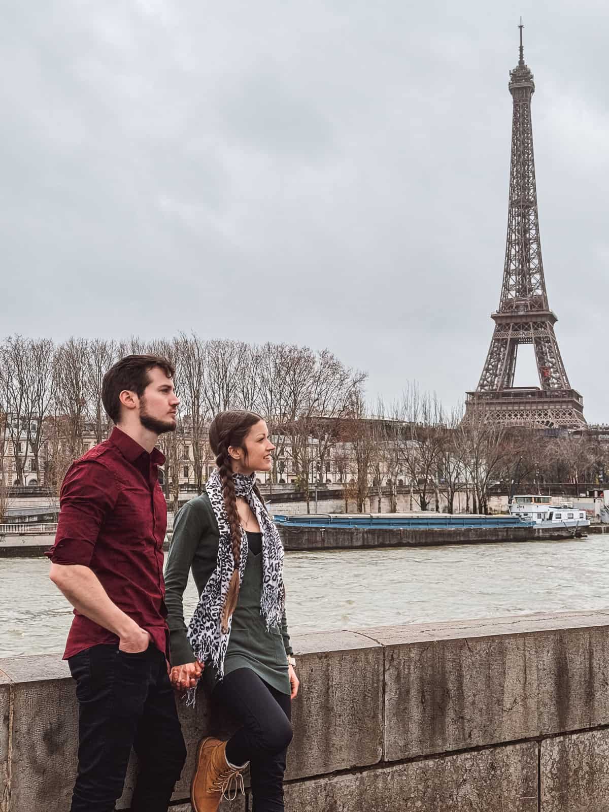 A man and woman, clad in casual attire, hold hands while looking out over the Seine River with the Eiffel Tower in the distant Paris skyline.