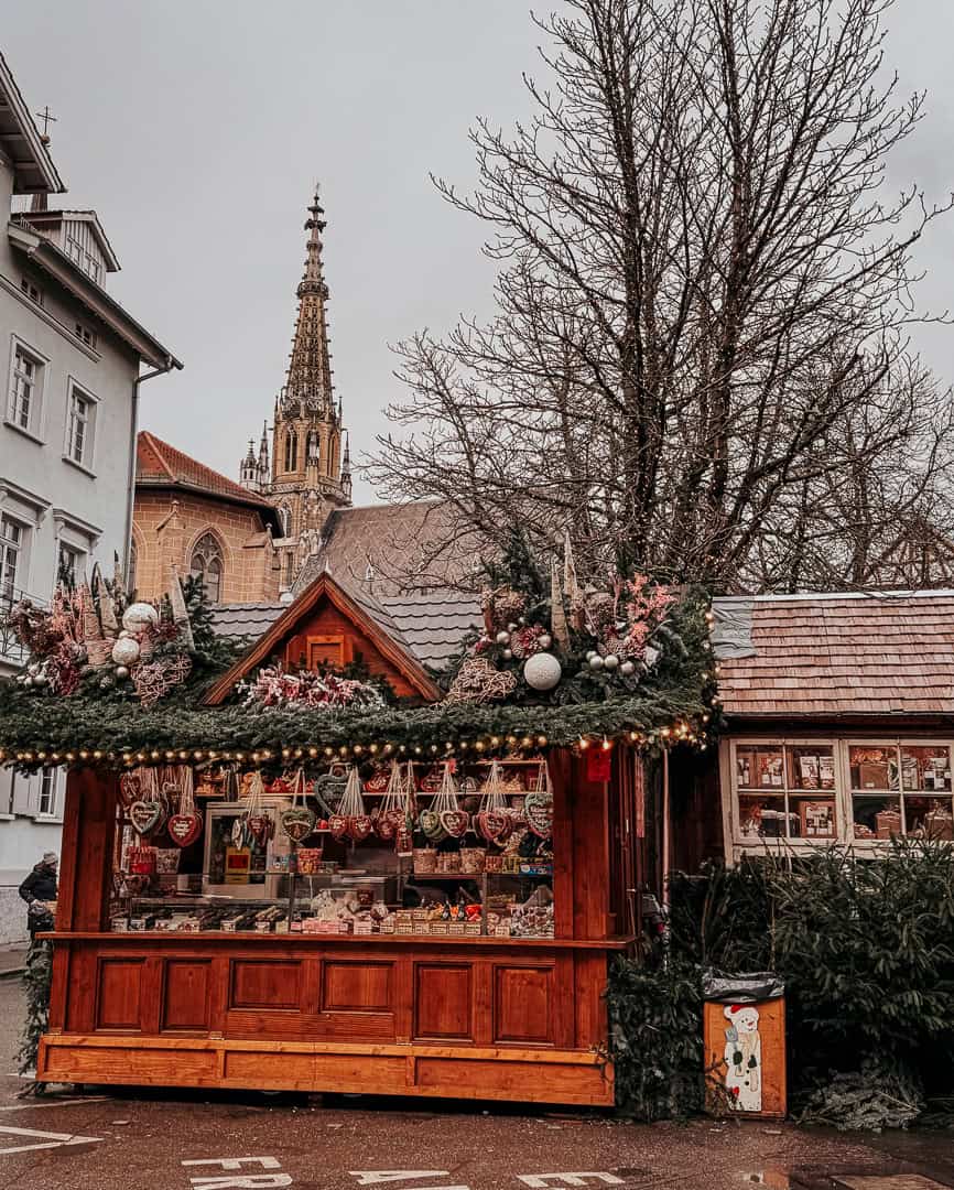 A quaint Christmas market stall in Esslingen, decorated with greenery and pink ornaments, offering a variety of sweets and treats with a gothic church spire in the background