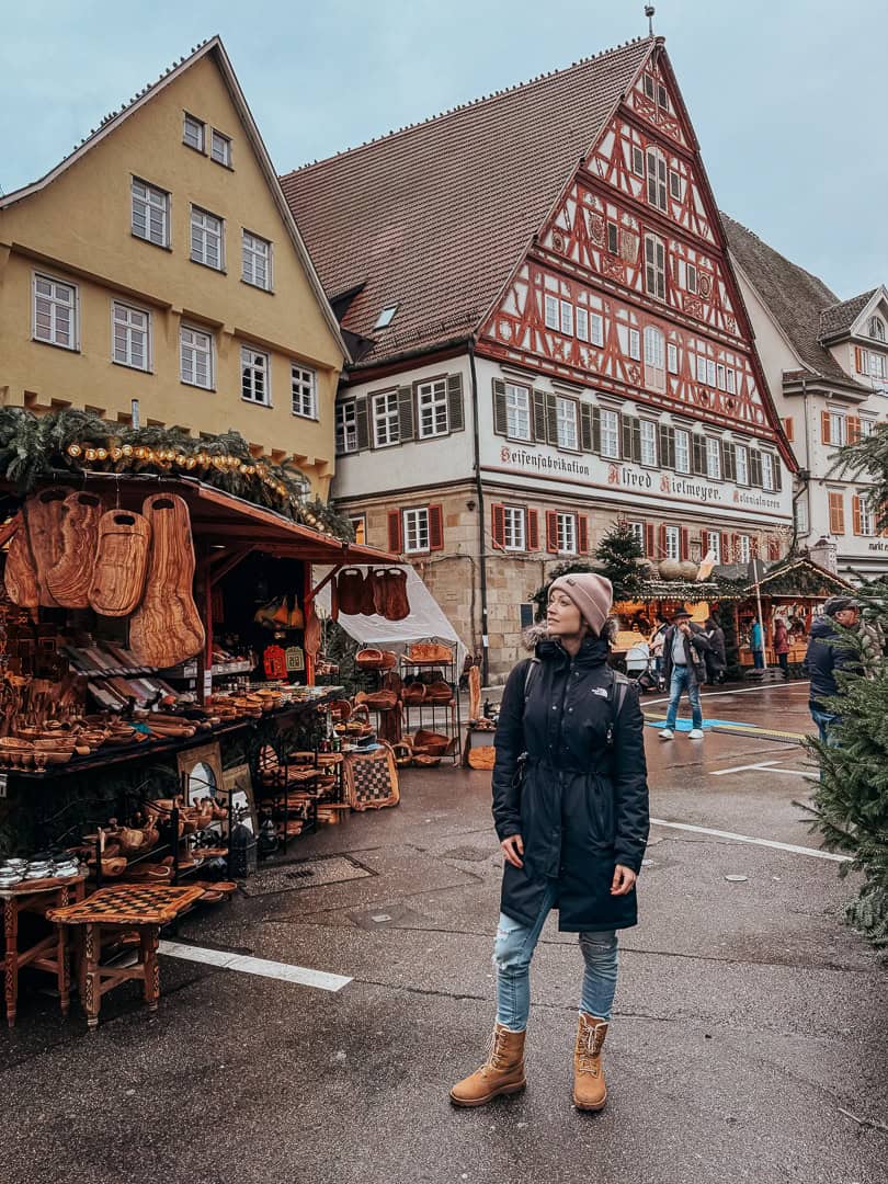 A person stands contemplatively in front of a traditional German Christmas market, with wooden stalls selling handcrafted goods, against the backdrop of a striking half-timbered building in Esslingen