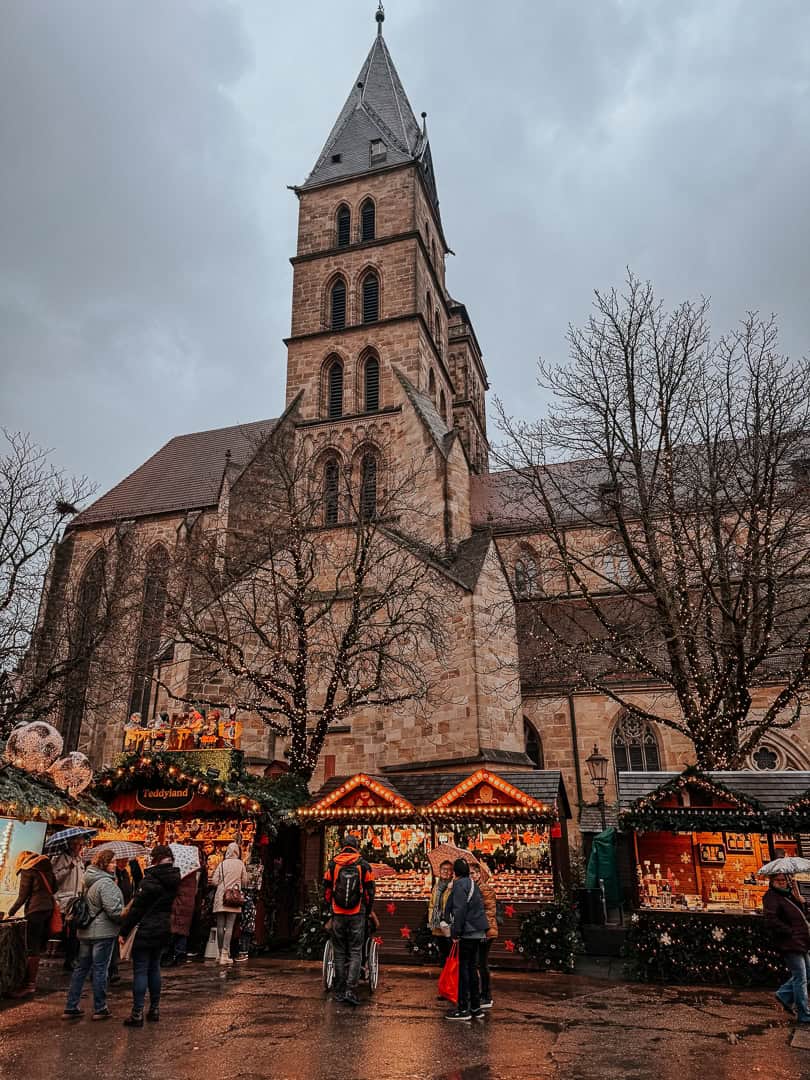Gothic church tower overlooking a bustling Christmas market with lit-up stalls and holiday shoppers in Esslingen am Neckar