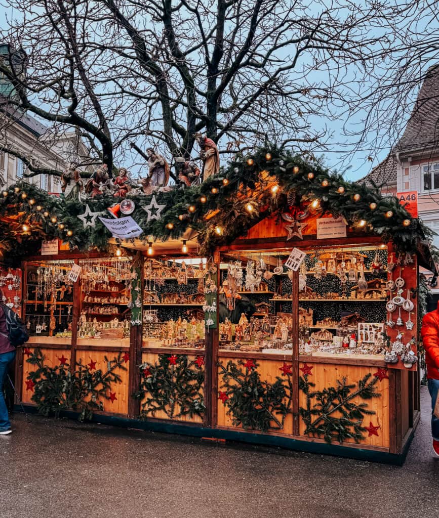 Shoppers explore a Christmas market booth in Colmar, topped with a nativity scene and draped in festive garlands, offering a selection of intricate ornaments.