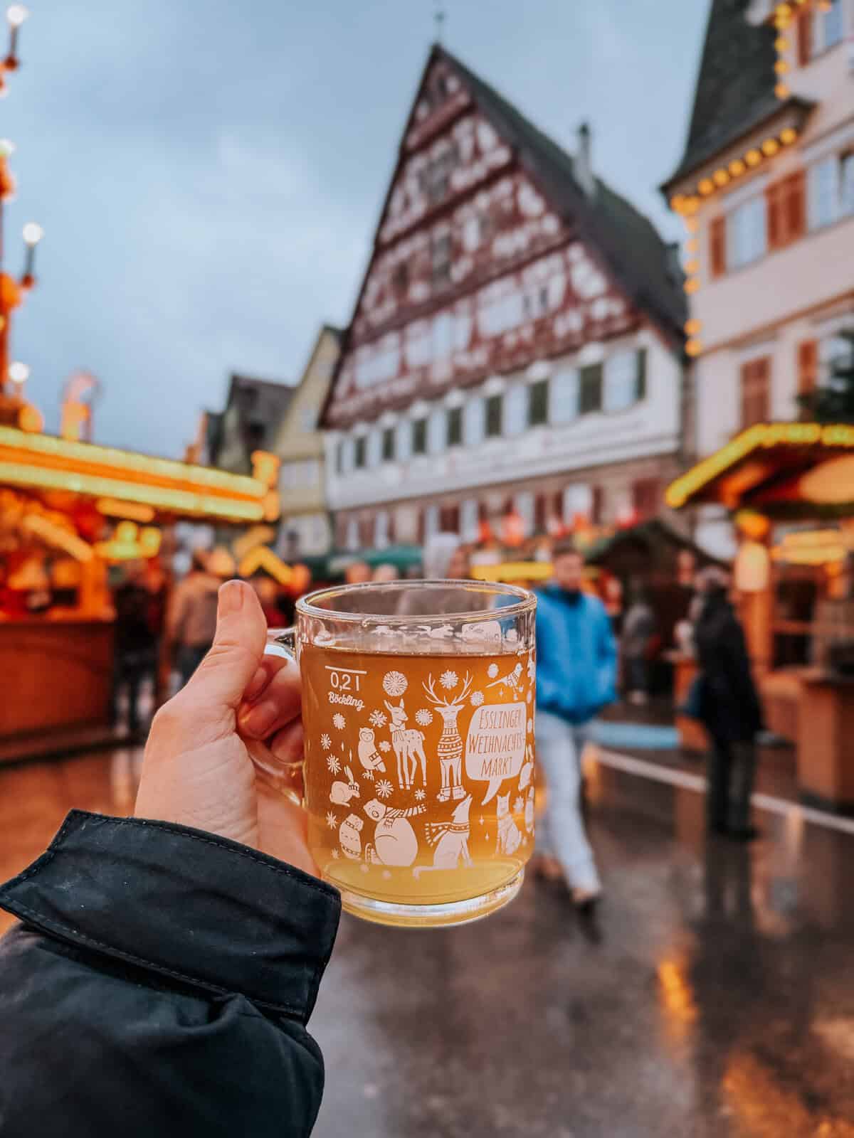Close-up of a hand holding a glass mug filled with steaming Glühwein at the Esslingen Christmas Market, with festive illustrations and market name on the cup
