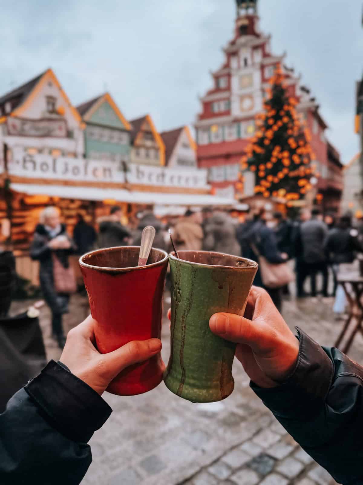 Two hands toasting with colorful ceramic mugs against the backdrop of a bustling Christmas market in Esslingen, with a beautifully lit half-timbered house.