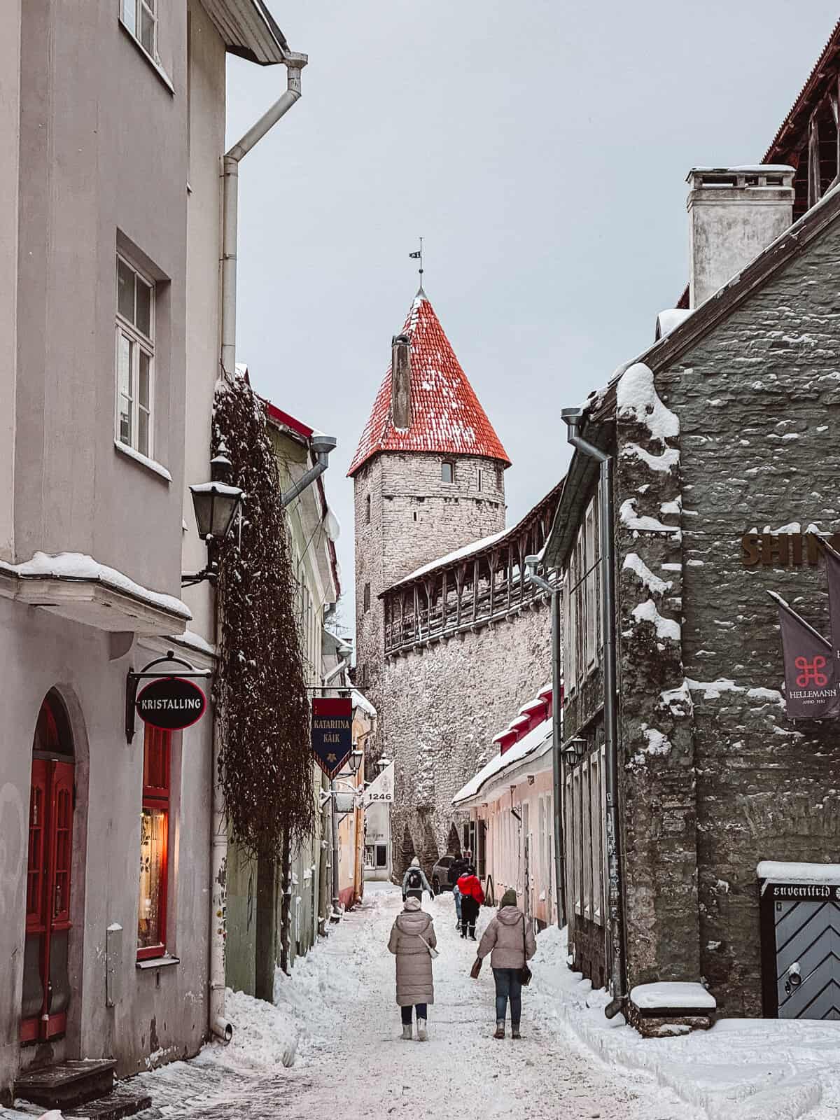 A snowy street in Tallinn's old town, lined with colorful buildings and people walking, evoking the charm of a European winter in Estonia, with the medieval town wall and its defensive tower completing the scene.