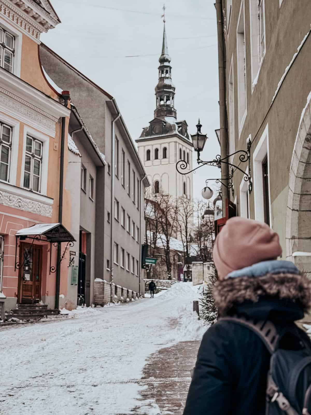a woman in a pink beanie looks up a snow street at a church in Tallinn
