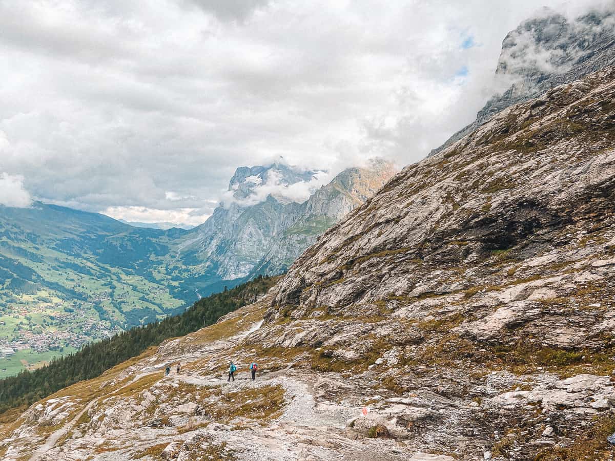 Hikers traversing a rocky alpine trail in the majestic Swiss mountains, with a sweeping view of the valley below and dramatic peaks shrouded by clouds, capturing the essence of adventure in the Swiss Alps.