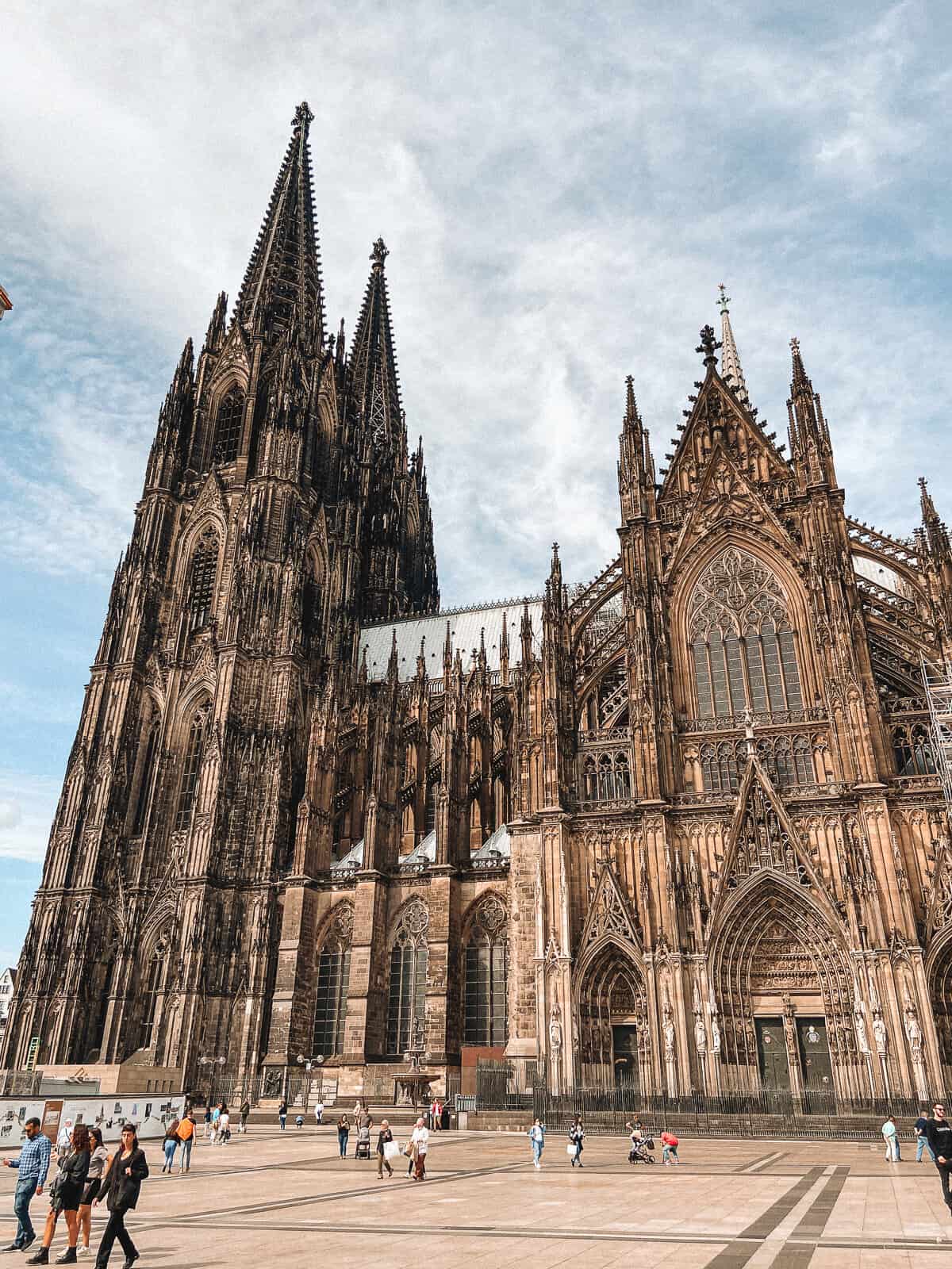 The imposing Gothic architecture of Cologne Cathedral stands grandly under a blue sky, with its intricate facades and towering spires, as tourists and locals alike roam the square in the foreground.
