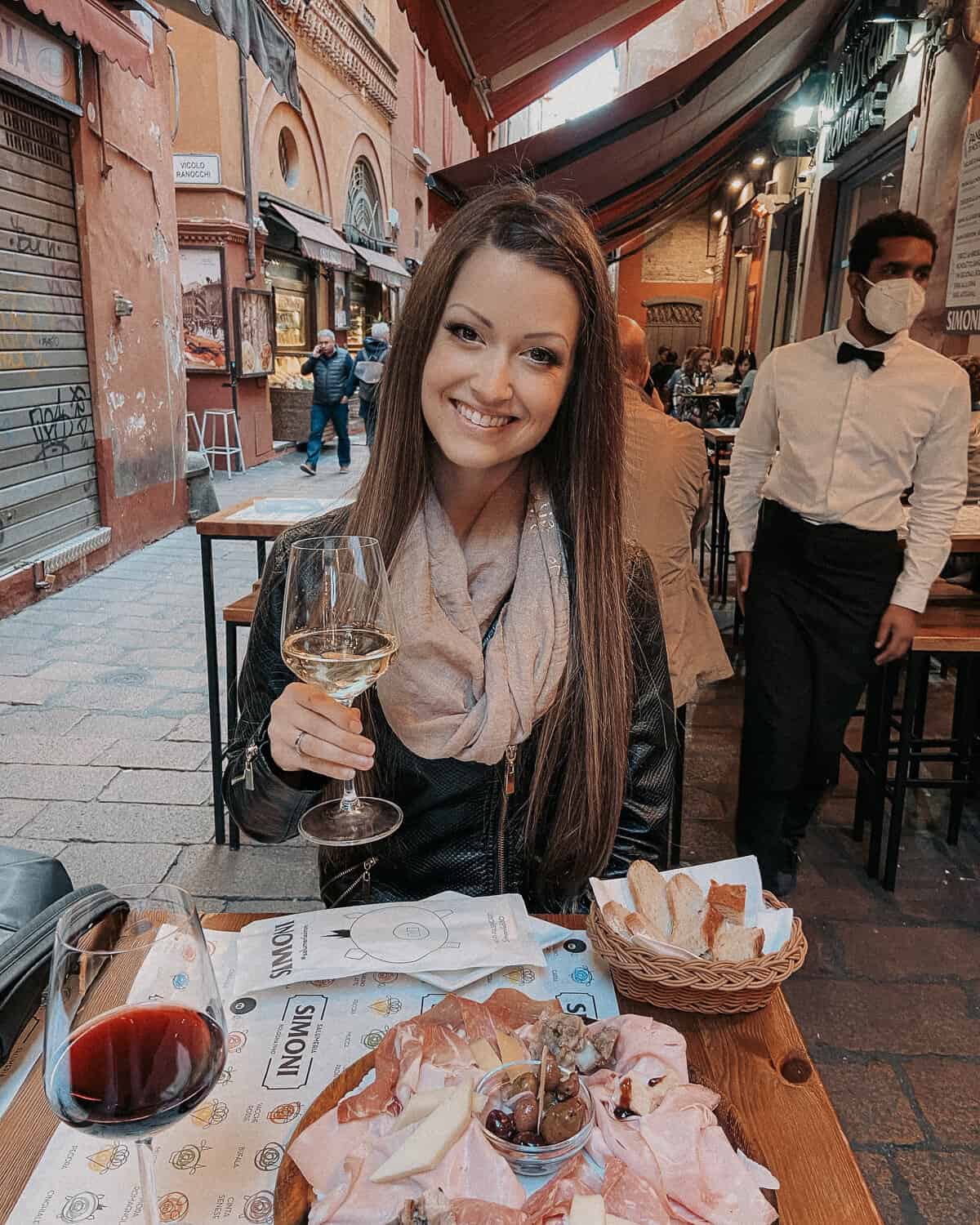A woman seated outdoors at a European bistro, smiling as she enjoys a glass of white wine and a platter of cured meats and olives, with the ambient hustle of the alley and a waiter in the background adding to the urban dining atmosphere.